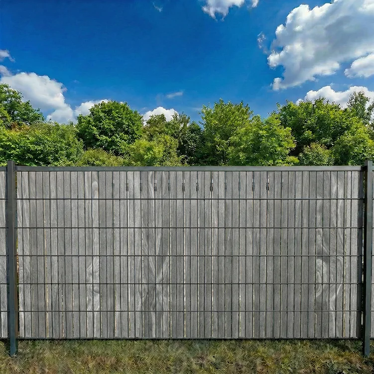 Accoya greyrfarbener Sichtschutzstreifen im Doppelstabmattenzaun als Sichtschutz im Garten bei blauem Himmel und Bäumen.