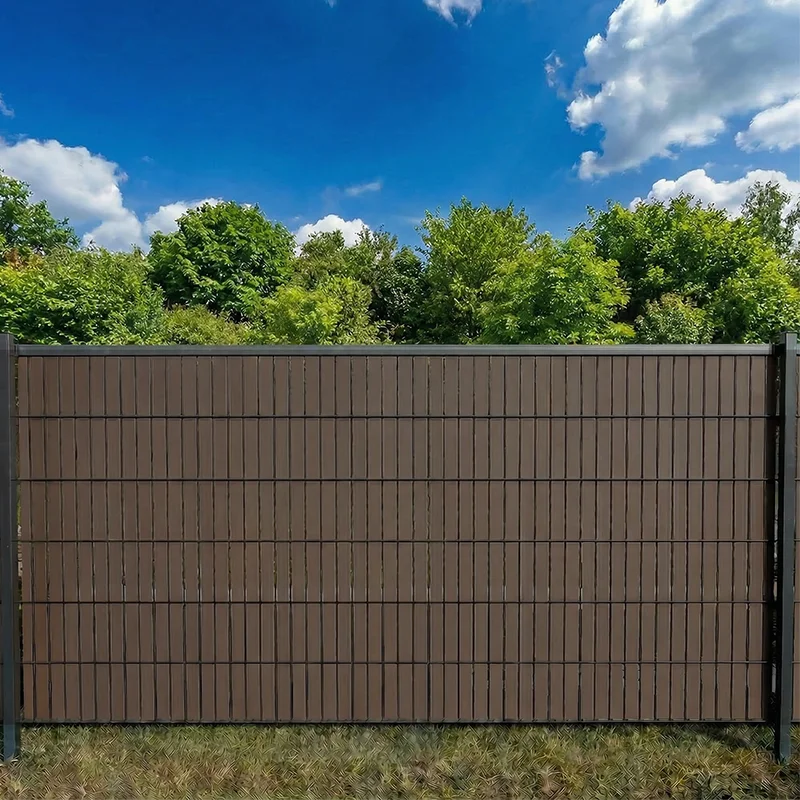 Flexio Copperfarbener Sichtschutzstreifen im Doppelstabmattenzaun als Sichtschutz im Garten bei blauem Himmel und Bäumen.