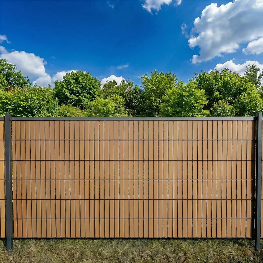 Flexio Copperfarbener Sichtschutzstreifen im Doppelstabmattenzaun als Sichtschutz im Garten bei blauem Himmel und Bäumen.